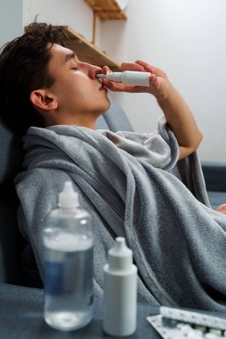 Adam burun spreyi kullanıyor ve hastalık tedavisi için ilaç ve ilaç alıyor. Adam üşüyor ve yatakta uzanıyor ve burnunu burun spreyiyle siliyor.
