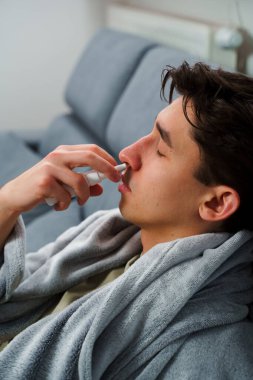 Adam burun spreyi kullanıyor ve hastalık tedavisi için ilaç ve ilaç alıyor. Adam üşüyor ve yatakta uzanıyor ve burnunu burun spreyiyle siliyor.
