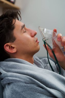 Battaniyeye sarılı hasta adam öksürük tedavisi için nebulizatörle solunum yapıyor. Hasta evde tedavi için astım spreyi kullanıyor.