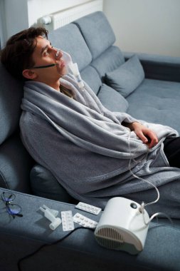 Battaniyeye sarılı hasta adam öksürük tedavisi için nebulizatörle solunum yapıyor. Hasta evde tedavi için astım spreyi kullanıyor.