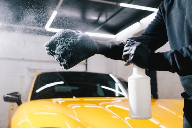 Adam detaylı işlemler sırasında araba seramik cilalaması için mikrofiber kumaşa balmumu püskürtür. Otomobil servisinde mikrofiberin üzerine mum spreyli araba temizliği.