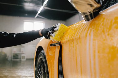 Oto yıkamadan önce sabun solüsyonu ve deterjanla araba yıkama prosedürü. İşçi, oto yıkamada sarı spor arabayı süngerle temizliyor.