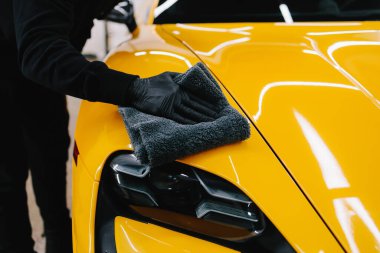 Tamirci, arabayı silmek için gri bir mikrofiber bez kullanıyor. Temizlikçi, sarı bir arabanın vücudunu mikrofiber bezle siler.