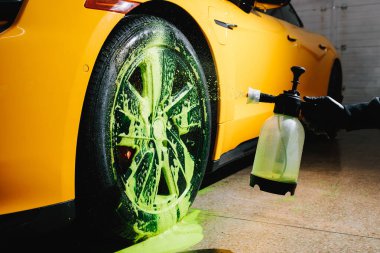 Sarı bir spor arabanın lastiğine sabun solüsyonu spreyi sıkarak yeşil deterjan sıkmak. Temizlikçi yeşil köpükle araba lastiklerini yıkıyor. Deterjanla fırça kullanıyor.