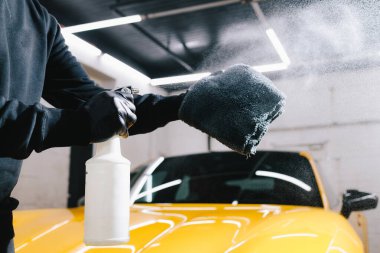 Adam detaylı işlemler sırasında araba seramik cilalaması için mikrofiber kumaşa balmumu püskürtür. Otomobil servisinde mikrofiberin üzerine mum spreyli araba temizliği.