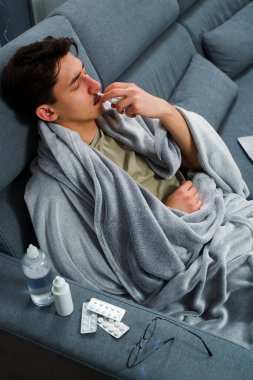 Adam üşümüş ve yatakta uzanıp burnunu burun spreyiyle siliyor. Adam burun spreyi kullanıyor ve hastalık tedavisi için ilaç ve ilaç alıyor.