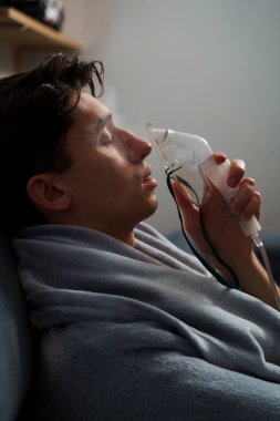 Hasta evde tedavi için nebulizer astım spreyi kullanıyor. Battaniyeye sarılı hasta adam öksürük tedavisi için nebulizatörle solunum yapıyor.