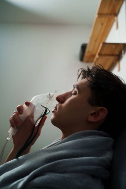Hasta evde tedavi için nebulizer astım spreyi kullanıyor. Battaniyeye sarılı hasta adam öksürük tedavisi için nebulizatörle solunum yapıyor.