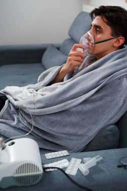 Hasta evde tedavi için nebulizer astım spreyi kullanıyor. Battaniyeye sarılı hasta adam öksürük tedavisi için nebulizatörle solunum yapıyor.