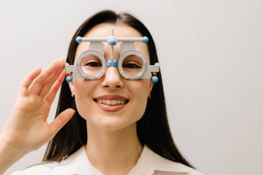 Kısa ya da uzun görüşlü bir kızın göz görme sistemini incelemek için deneme gözlüğü seçimi için oftalmolog muayenesi. Beyaz arka planda deneme çerçeveli gözlüklü kız