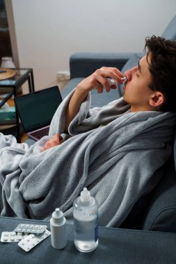 Adam üşümüş ve yatakta uzanıp burnunu burun spreyiyle siliyor. Adam burun spreyi kullanıyor ve hastalık tedavisi için ilaç ve ilaç alıyor.