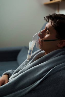 Hasta evde tedavi için nebulizer astım spreyi kullanıyor. Battaniyeye sarılı hasta adam öksürük tedavisi için nebulizatörle solunum yapıyor.