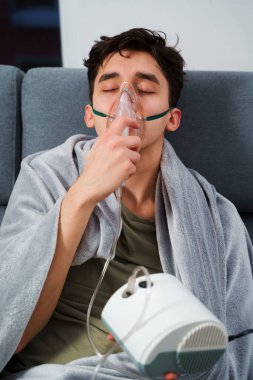 Battaniyeye sarılı hasta adam öksürük tedavisi için nebulizatörle solunum yapıyor. Hasta evde tedavi için astım spreyi kullanıyor.