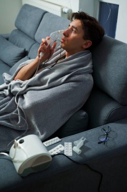 Hasta evde tedavi için nebulizer astım spreyi kullanıyor. Battaniyeye sarılı hasta adam öksürük tedavisi için nebulizatörle solunum yapıyor.