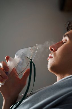 Evde hastalık tedavisi için kompresör nebulizatörü kullanılıyor. Öksürük tedavisi için oksijen maskesi takan bir adam.