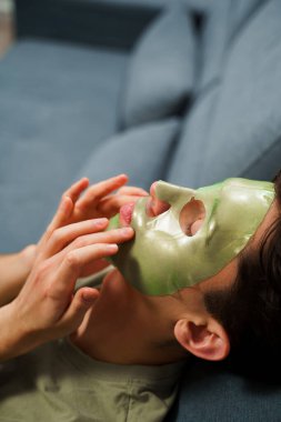 Genç adam susuzluk efektli yeşil yüz maskesi kullanıyor. Erkek güzellik prosedürleri. Derisini sulandırıcı ve nemlendirici bir maskeyle besleyip şımartıyor.