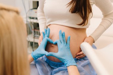 Mavi eldivenli jinekolog, tıbbi standartlara göre değerlendirmek için hamile karnı inceliyor. Jinekolog jinekoloji kliniğinde kadının karnında kalp oluşturur.