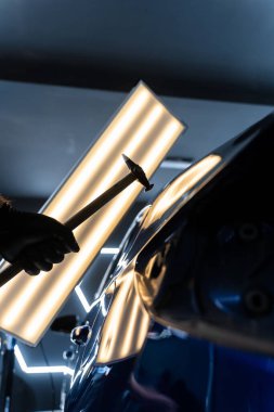 Ağrısız çukur onarma PDR hassasiyet için ışık yansıması altında ezilmeyi içerir. Teknisyen, acısız çukur onarımı sırasında arabanın kaputundaki göçükleri çıkarmak için PDR araçları kullanıyor.