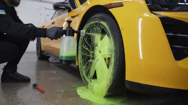 Adam araba yıkama videosunda tekerlek lastiğine sabun köpüğü sıkıyor. İşçi, sarı bir arabanın lastiğini sabun solüsyonuyla temizliyor.