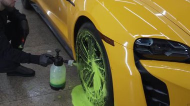 İşçi 4K videosu sırasında sabun solüsyonlu sarı bir arabanın lastiğini temizliyor. Adam oto yıkamada lastiğe sabun köpüğü sıkıyor.