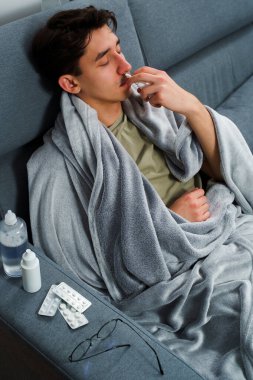 Adam üşümüş ve yatakta uzanıp burnunu burun spreyiyle siliyor. Adam burun spreyi kullanıyor ve hastalık tedavisi için ilaç ve ilaç alıyor.