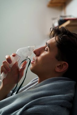 Hasta evde tedavi için nebulizer astım spreyi kullanıyor. Battaniyeye sarılı hasta adam öksürük tedavisi için nebulizatörle solunum yapıyor.