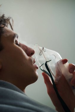 Hasta evde tedavi için nebulizer astım spreyi kullanıyor. Battaniyeye sarılı hasta adam öksürük tedavisi için nebulizatörle solunum yapıyor.