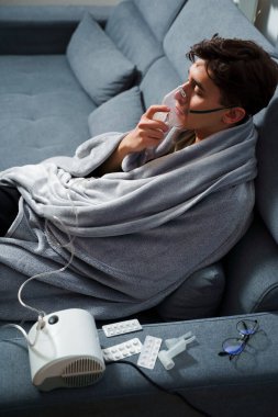 Hasta evde tedavi için nebulizer astım spreyi kullanıyor. Battaniyeye sarılı hasta adam öksürük tedavisi için nebulizatörle solunum yapıyor.