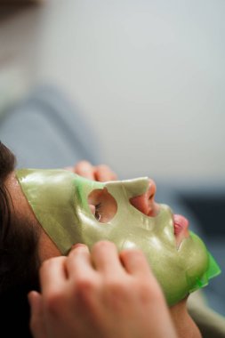 Genç adam susuzluk efektli yeşil yüz maskesi kullanıyor. Erkek güzellik prosedürleri. Derisini sulandırıcı ve nemlendirici bir maskeyle besleyip şımartıyor.