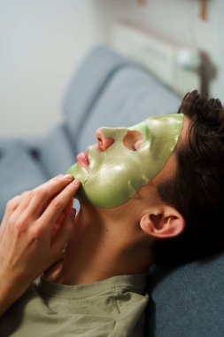 Genç adam susuzluk efektli yeşil yüz maskesi kullanıyor. Erkek güzellik prosedürleri. Derisini sulandırıcı ve nemlendirici bir maskeyle besleyip şımartıyor.