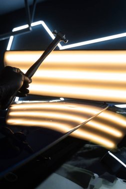 PDR Acısız araba göçüğü temizleme. PDR teknisyeni, kaputtaki çukur düzeltmesini kontrol etmek için yansıma lambasına özel bir çekiç tutuyor.