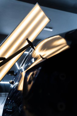 Teknisyen, acısız çukur onarımı sırasında arabanın kaputundaki göçükleri çıkarmak için PDR aletleri kullanıyor. Ağrısız çukur onarma PDR hassasiyet için ışık yansıması altında ezilmeyi içerir.