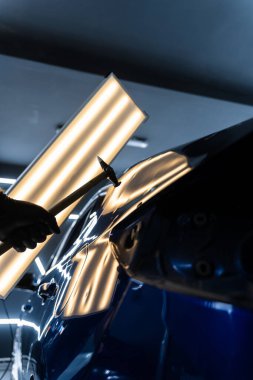 Teknisyen, acısız çukur onarımı sırasında arabanın kaputundaki göçükleri çıkarmak için PDR aletleri kullanıyor. Ağrısız çukur onarma PDR hassasiyet için ışık yansıması altında ezilmeyi içerir.