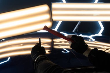 Teknisyen, acısız çukur onarımı sırasında arabanın kaputundaki göçükleri çıkarmak için PDR aletleri kullanıyor. Ağrısız çukur onarımı (PDR) hassasiyet için ışık yansıması altında ezilmeyi içerir