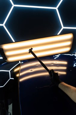 PDR Acısız araba göçüğü temizleme. PDR teknisyeni, kaputtaki çukur düzeltmesini kontrol etmek için yansıma lambasına özel bir çekiç tutuyor.