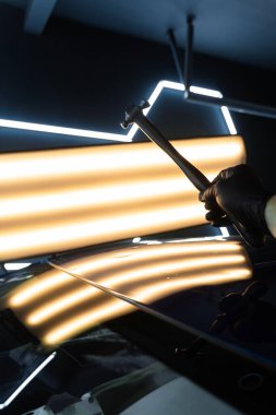 PDR teknisyeni, kaputtaki çukur düzeltmesini kontrol etmek için yansıma lambasına özel bir çekiç tutuyor. Acısız bir araba göçüğü.