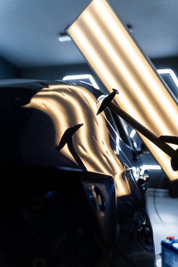 Ağrısız çukur onarma PDR hassasiyet için ışık yansıması altında ezilmeyi içerir. Teknisyen, acısız çukur onarımı sırasında arabanın kaputundaki göçükleri çıkarmak için PDR araçları kullanıyor.