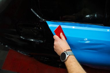 Araç ambalajı, pürüzsüz ve kırışıklıksız yapışma için cam sileceği darbelerini içeriyor. Ayrıntılı uzman, araba yüzeyinde mavi PPF ambalajını düzleştirmek için kırmızı cam ambalajı kullanır