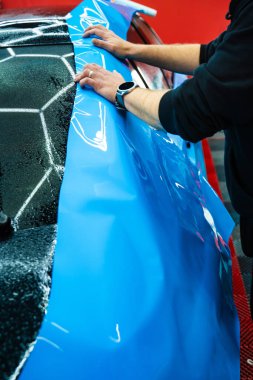 Tüm detaylar sırasında arabaya PPF ambalajı uygulanıyor. Detaylandırma merkezine kalın mavi vinil ambalaj kurulumu. Detaylar hatları sığdırmak için ustalıkla paket uygular ve pürüzsüz, pürüzsüz paket hizalamasını sağlar