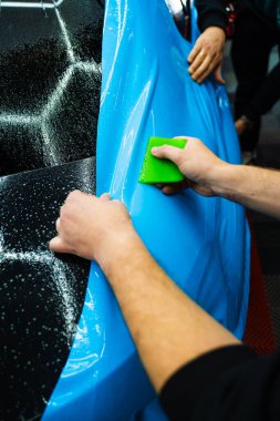 Mavi PPF, yeşil cam sileceği ile arabayı temizlerken ve hasarı engellemek için uygulanmış. Yeşil bir cam sileceği kullanarak yüksek parlak mavi bir PPF uygulamak, arabanın baloncuksuz ve pürüzsüz bir yüzey sağlamasını sağlar