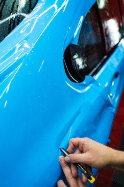 Mavi PPF 'yi bıçakla kesiyorum. Detaylı iş sırasında mavi boya koruma filmini bıçakla kesmek hassas kenarlar sağlar. Detaylı teknisyen mükemmel kontur için bir araba paneli üzerindeki mavi PPF buduyor