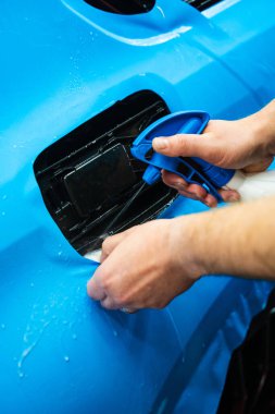 Detaylı uzmanlar mavi PPF filminin kontrollü uygulaması için hidrasyon spreyi kullanırlar. Detaylayıcı mükemmel, kabarcıksız hizalama için canlı mavi vinil sarma uygulamak için sprey destekli yöntem kullanır