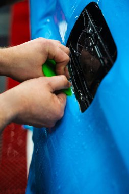 Mavi PPF, yeşil cam sileceği ile arabayı temizlerken ve hasarı engellemek için uygulanmış. Yeşil bir cam sileceği kullanarak yüksek parlak mavi bir PPF uygulamak, arabanın baloncuksuz ve pürüzsüz bir yüzey sağlamasını sağlar