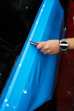 Araç ambalajı, pürüzsüz ve kırışıklıksız yapışma için cam sileceği darbelerini içeriyor. Ayrıntılı uzman, araba yüzeyinde mavi PPF ambalajını düzleştirmek için kırmızı cam ambalajı kullanır