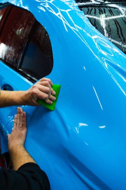 Mavi PPF, yeşil cam sileceği ile arabayı temizlerken ve hasarı engellemek için uygulanmış. Yeşil bir cam sileceği kullanarak yüksek parlak mavi bir PPF uygulamak, arabanın baloncuksuz ve pürüzsüz bir yüzey sağlamasını sağlar