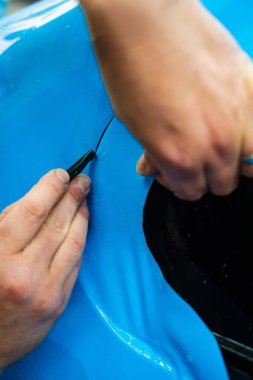 Mavi PPF 'yi bıçakla kesiyorum. Detaylı iş sırasında mavi boya koruma filmini bıçakla kesmek hassas kenarlar sağlar. Detaylı teknisyen mükemmel kontur için bir araba paneli üzerindeki mavi PPF buduyor