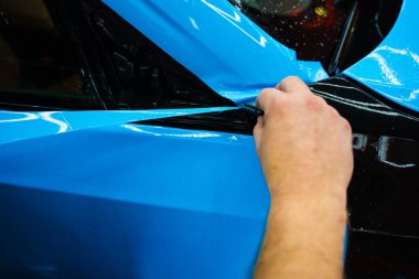 Mavi PPF 'yi bıçakla kesiyorum. Detaylı iş sırasında mavi boya koruma filmini bıçakla kesmek hassas kenarlar sağlar. Detaylı teknisyen mükemmel kontur için bir araba paneli üzerindeki mavi PPF buduyor