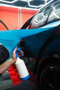 Detaylı uzmanlar mavi PPF filminin kontrollü uygulaması için hidrasyon spreyi kullanırlar. Detaylayıcı mükemmel, kabarcıksız hizalama için canlı mavi vinil sarma uygulamak için sprey destekli yöntem kullanır