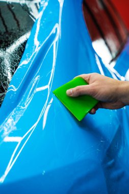 Mavi PPF, yeşil cam sileceği ile arabayı temizlerken ve hasarı engellemek için uygulanmış. Yeşil bir cam sileceği kullanarak yüksek parlak mavi bir PPF uygulamak, arabanın baloncuksuz ve pürüzsüz bir yüzey sağlamasını sağlar