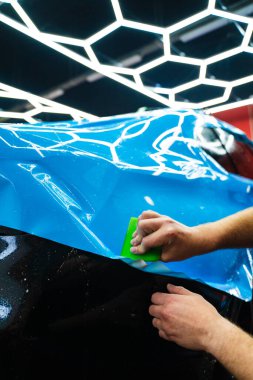 Mavi PPF, yeşil cam sileceği ile arabayı temizlerken ve hasarı engellemek için uygulanmış. Yeşil bir cam sileceği kullanarak yüksek parlak mavi bir PPF uygulamak, arabanın baloncuksuz ve pürüzsüz bir yüzey sağlamasını sağlar